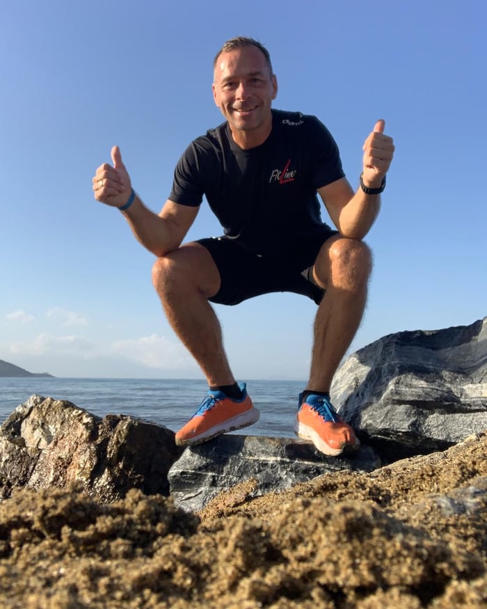 Michael Wehmann in tiefer Hocke auf Felsen am Meer, im schwarzen FitLine-Shirt, beide Daumen nach oben.