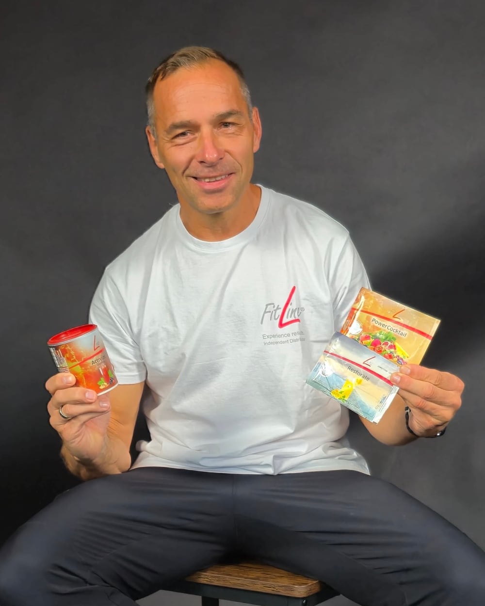 Michael Wehmann sitzt auf einem Hocker im weißen FitLine-Shirt und hält eine Dose Activize und die Beutel PowerCocktail und Restorate in den Händen.
